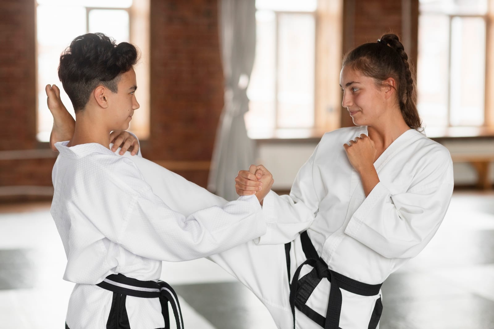 Two martial artists in white uniforms with black belts practising a close-range block and grab in a bright training hall